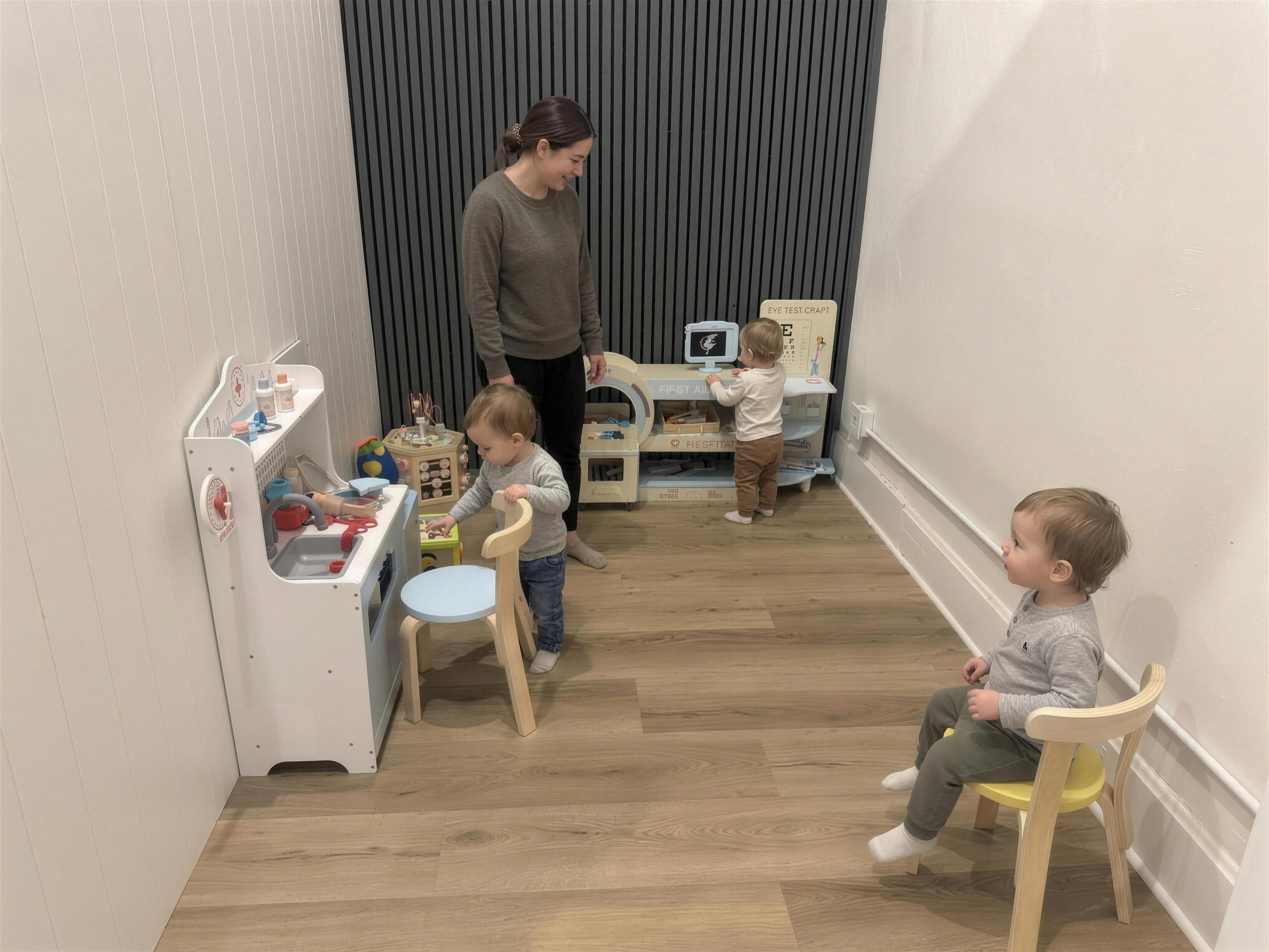 A grown-up and toddlers playing at the little clinic, with its first-aid cart and eye chart.