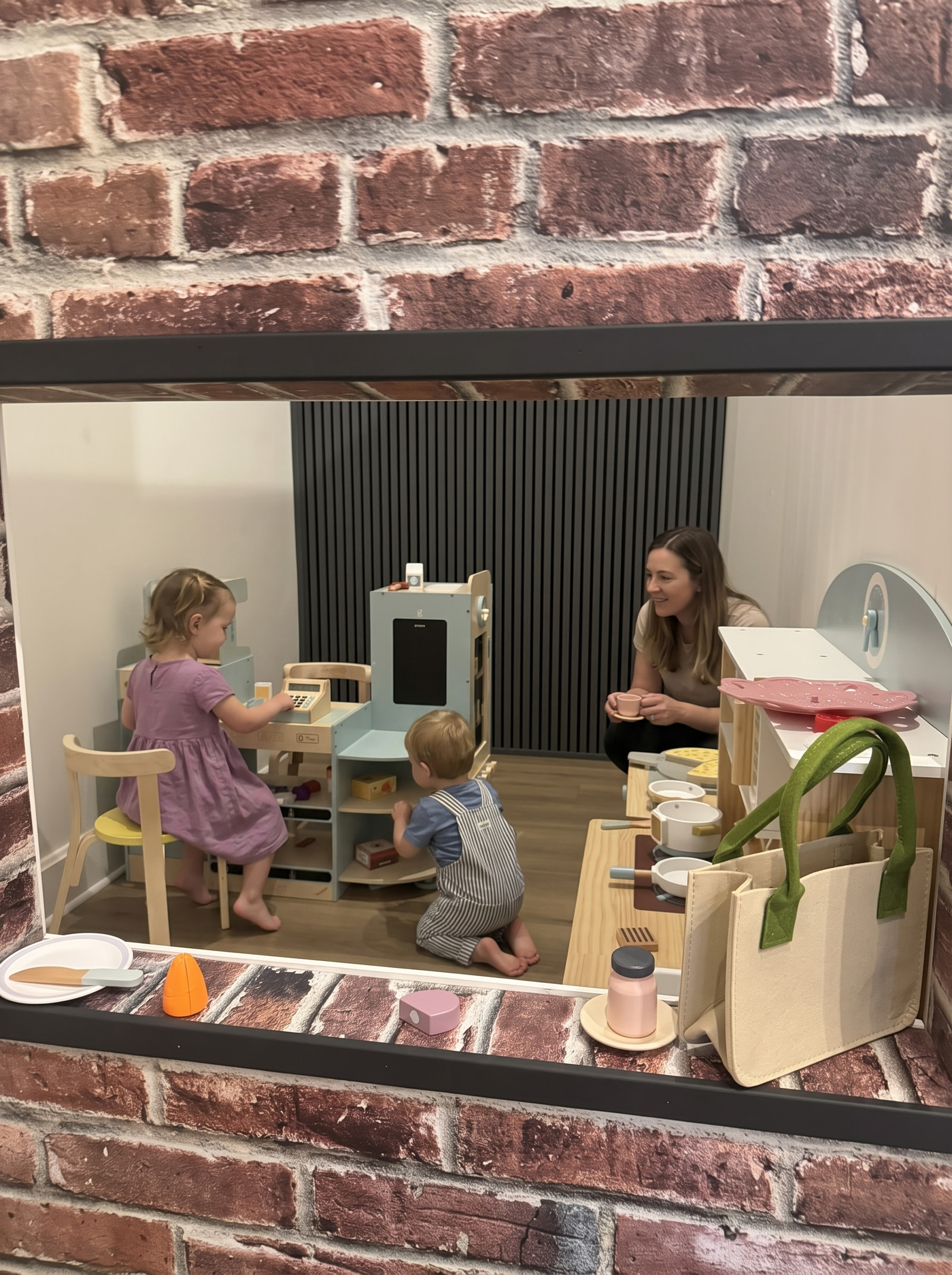 A grown-up and two children at a pretend tea party in the play kitchen, seen through the brick window.