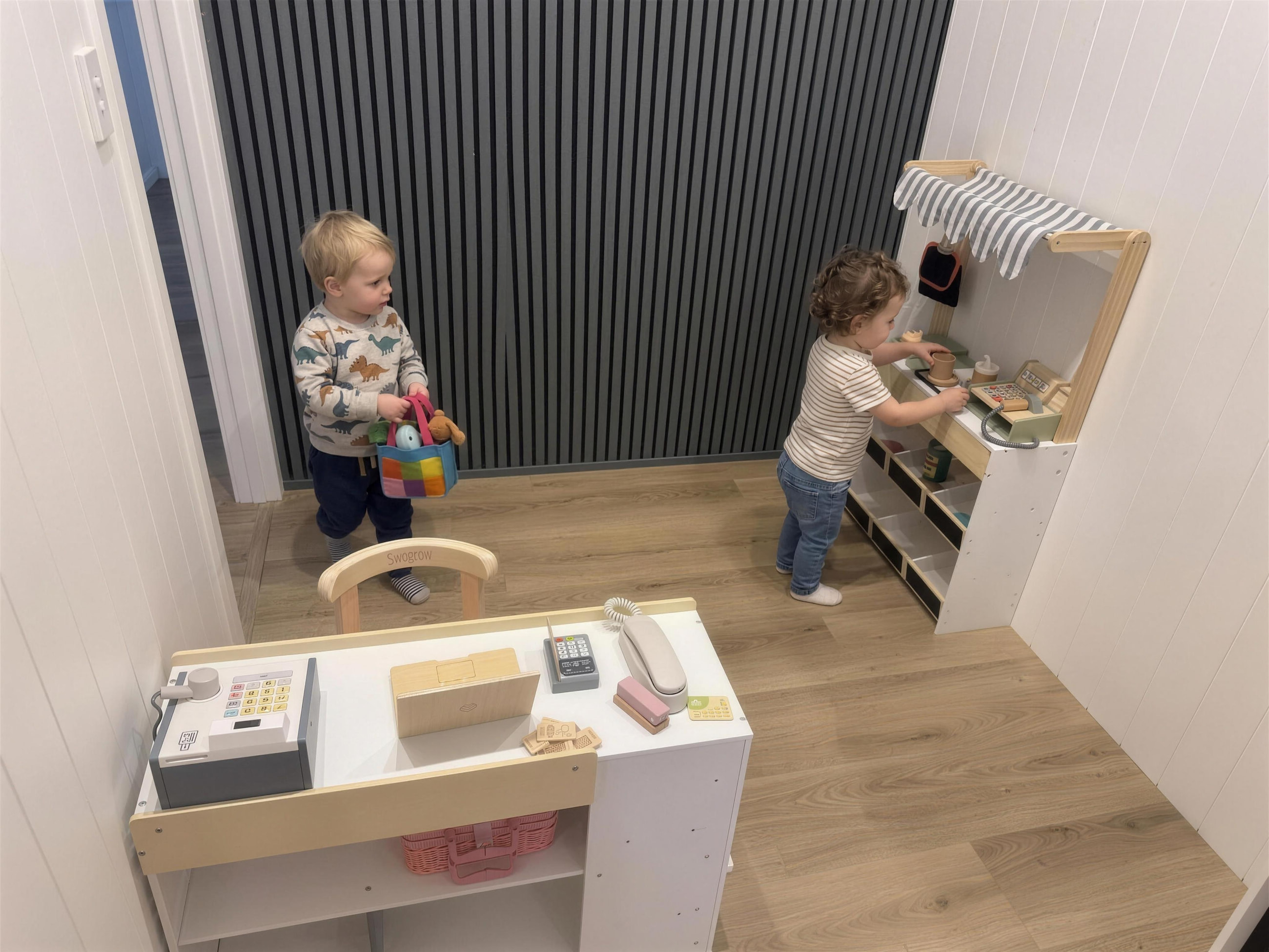 A toddler carrying a shopping basket past the little market stall.