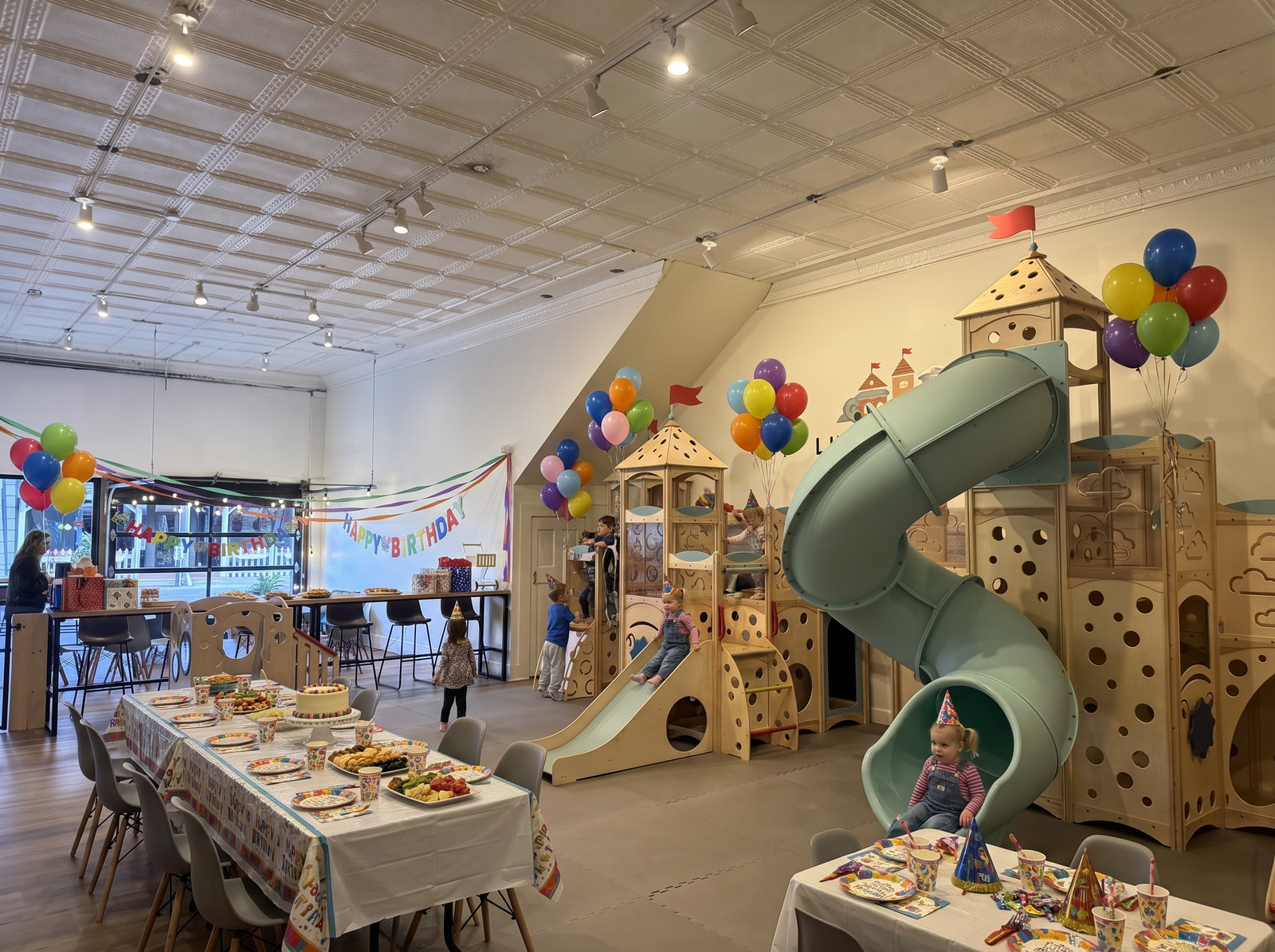 Little Town set up for a birthday party — balloons on the castle, a Happy Birthday banner, and long tables laid with cake and snacks while kids play on the slide.
